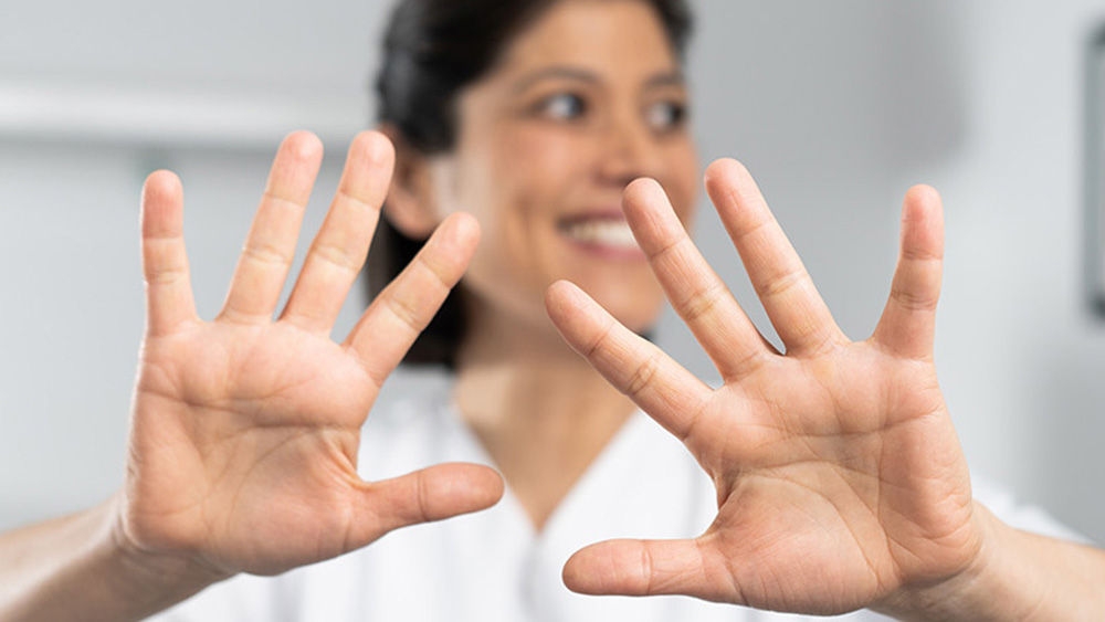 A person is wearing a white lab coat and is facing the camera. Both hands are stretched forward with fingers spread apart. The person's face is intentionally blurred to protect their identity. In the background, there is a blurred indoor setting with a picture hanging on the wall and a piece of furniture that looks like a bed or a couch.