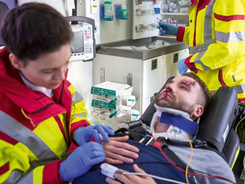 A female healthcare professional is performing first aid on a man with blood on his face inside an ambulance, using medical pumps