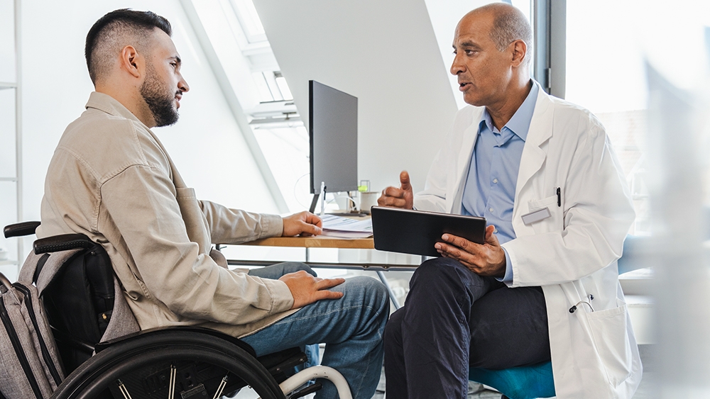 A patient in a wheelchair sits in front of a doctor who explains something to him with an ipad in his hand