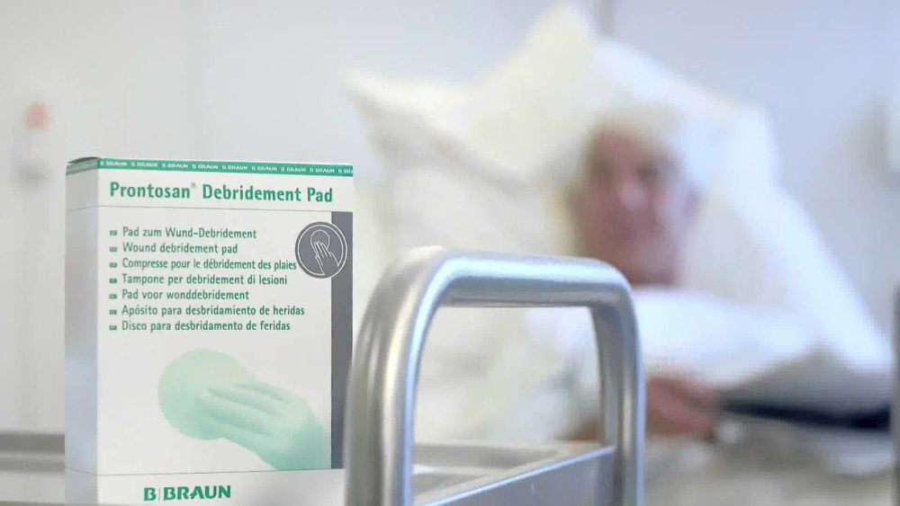 a pack of Prontosan® Debridement Pad picktured in the hospital room with the patient lying on the bed in the background