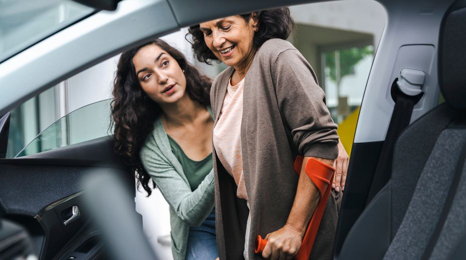 A patient using a crutch and assisted by another individual in getting into their vehicle.