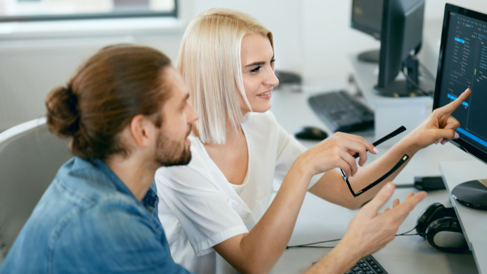 Two Health care worker are sitting in front of a PC. She shows him something on the screen.