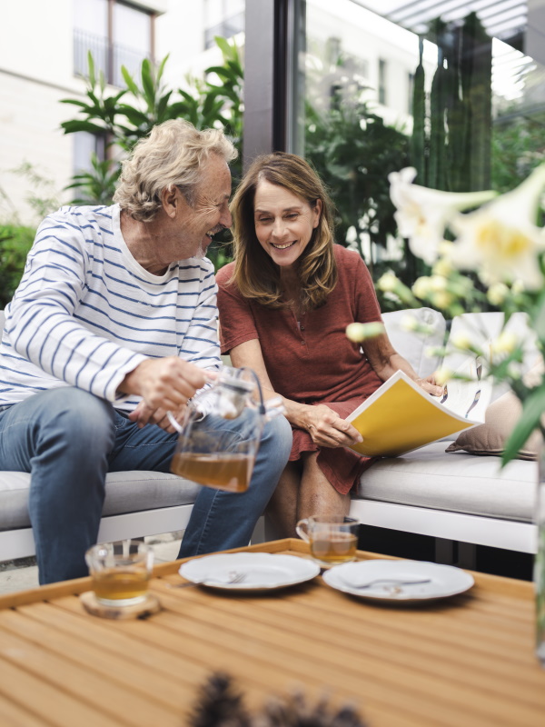 A man with a teapot and a woman with a magazine sit on a sofa and laugh