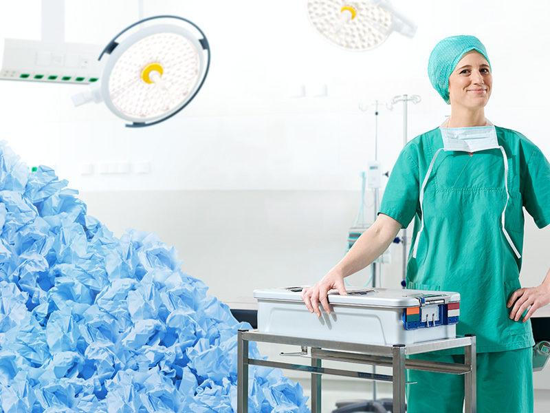 A female healthcare professional stands behind a table with a large pile of used medical clothing in the background