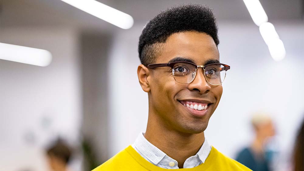 Close-up of a smiling individual with a yellow top and glasses standing in a bright setting. 