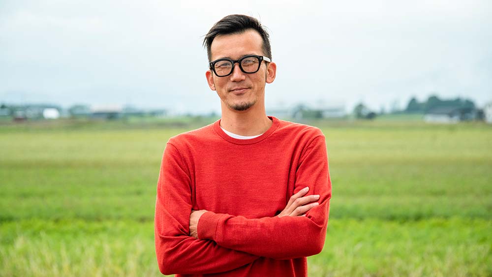 A person wearing glasses stands confidently and with crossed arms in a vast field.
